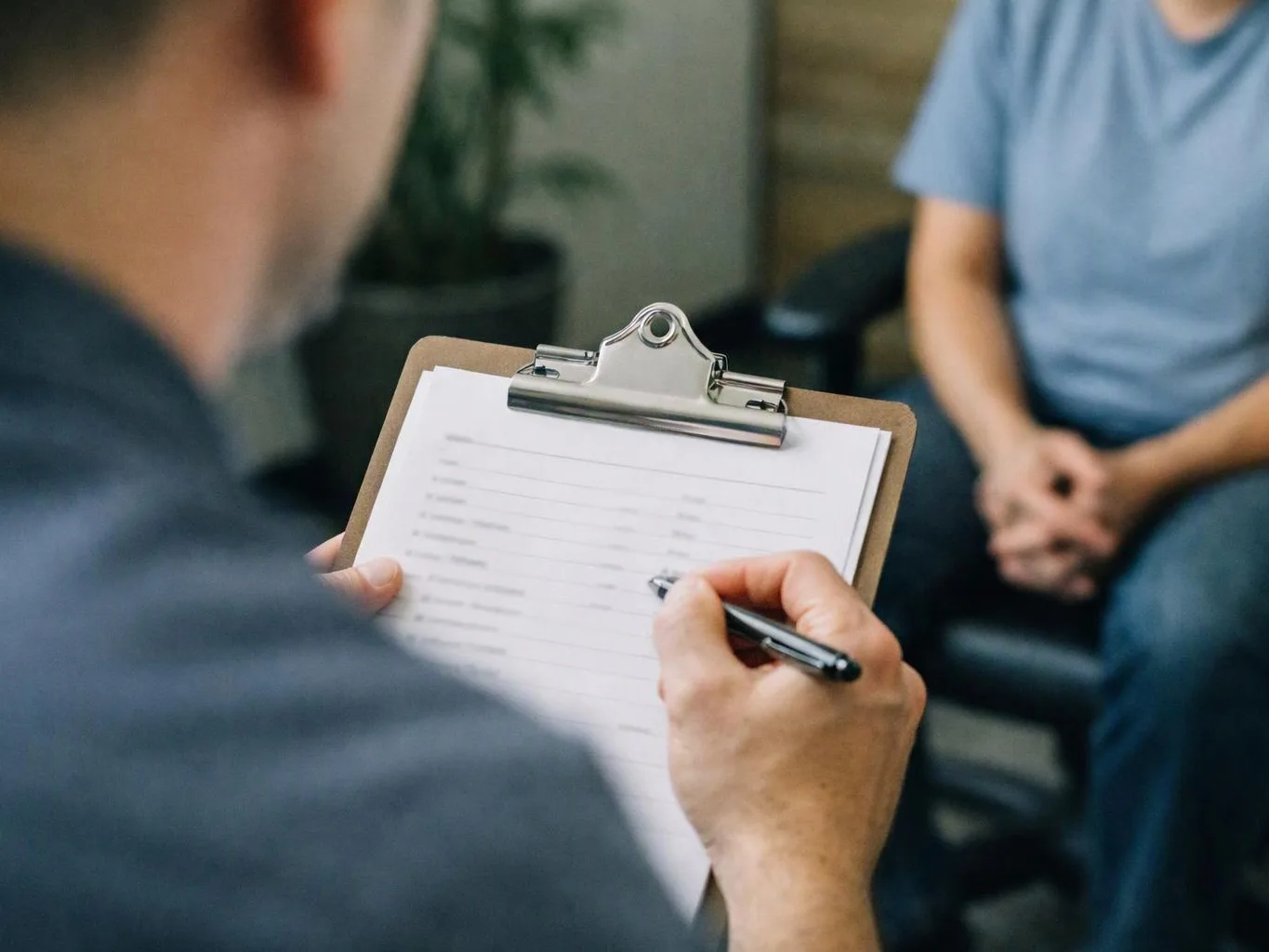 A person writes on a clipboard while another sits across from them in a waiting room