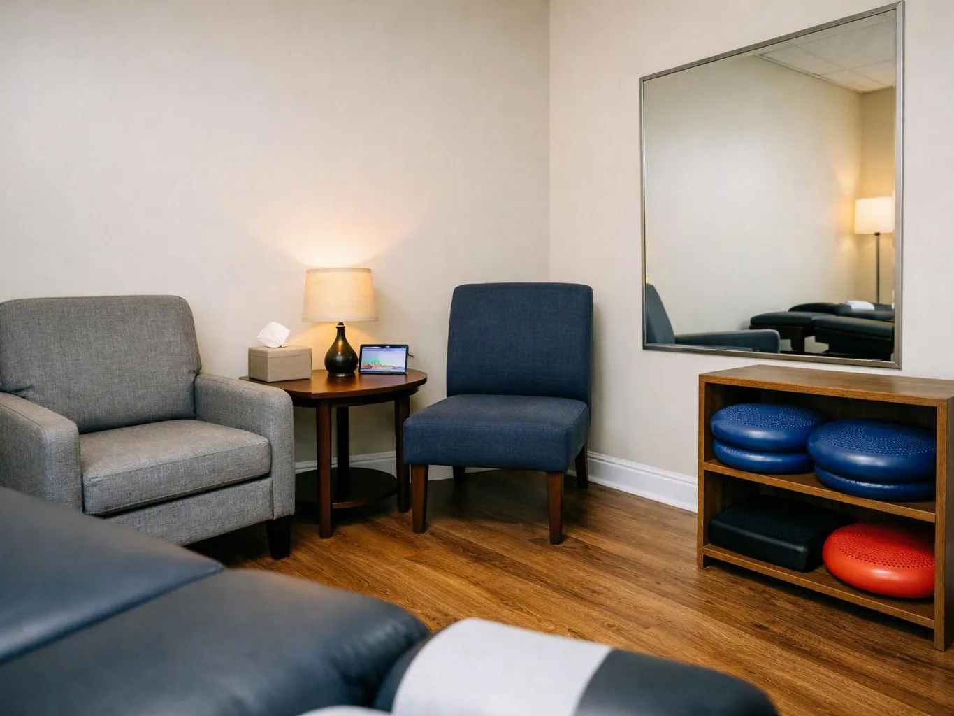 A quiet treatment room holds a gray chair, blue chair, lamp table, mirror, and exercise cushions on a shelf