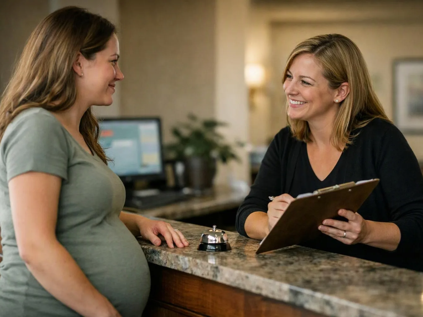 Two women smile across a reception counter, one holding a clipboard while the other stands with a pregnant belly